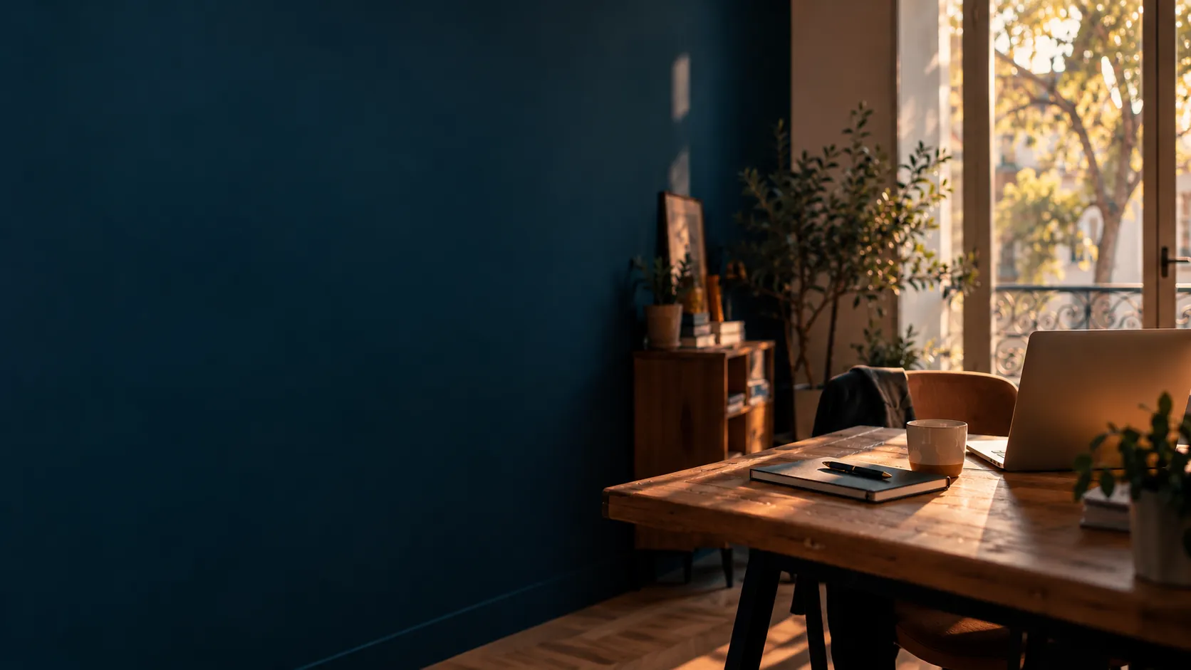 Un bureau en bois baigné de lumière devant un mur bleu profond, avec un carnet, une tasse et un ordinateur portable